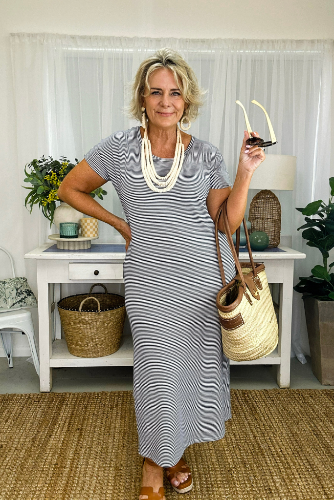A woman wearing a navy and white striped maxi dress with a scoop neckline, short sleeves, and side splits to the knee. She is accessorized with multiple necklaces, a bracelet, and carrying a straw bag.