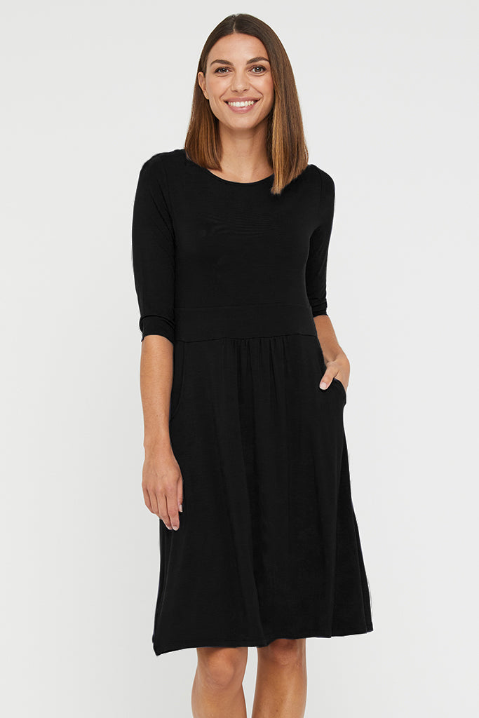 A woman wearing a knee-length black dress with 3/4 sleeves, a round neckline, and in-built waistband with pleat detail. The dress features two side pockets and is pictured in a neutral setting.