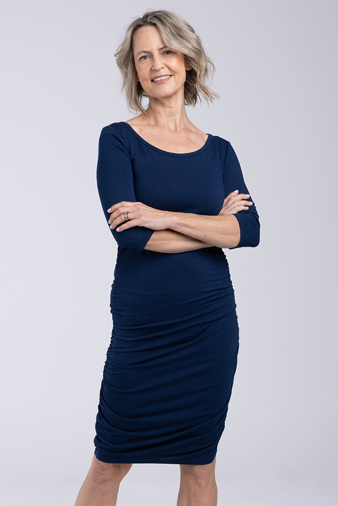 A navy-colored, 3/4 sleeve ruched dress on a model who is standing with her arms crossed. The dress features a scoop neckline and ruching down both sides, with a length that sits above the knee.