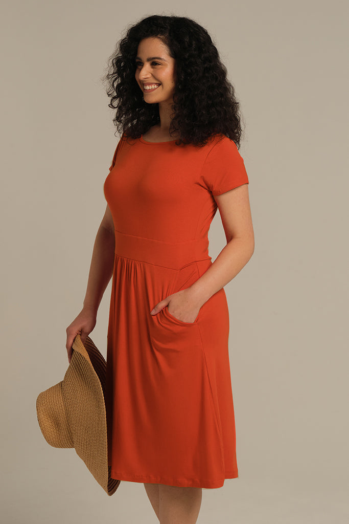 A woman wearing a knee-length orange dress with a round neckline, short sleeves, and pleat details at the waist. She is holding a hat and has her hand in a pocket.