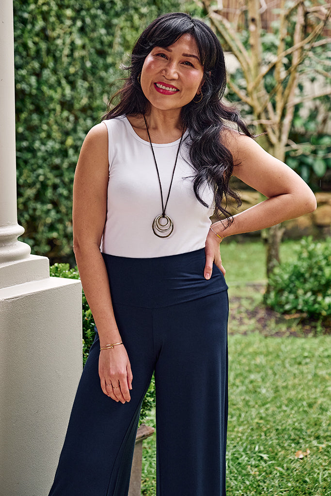 Woman in white sleeveless tank top and navy pants standing outdoors with greenery in the background