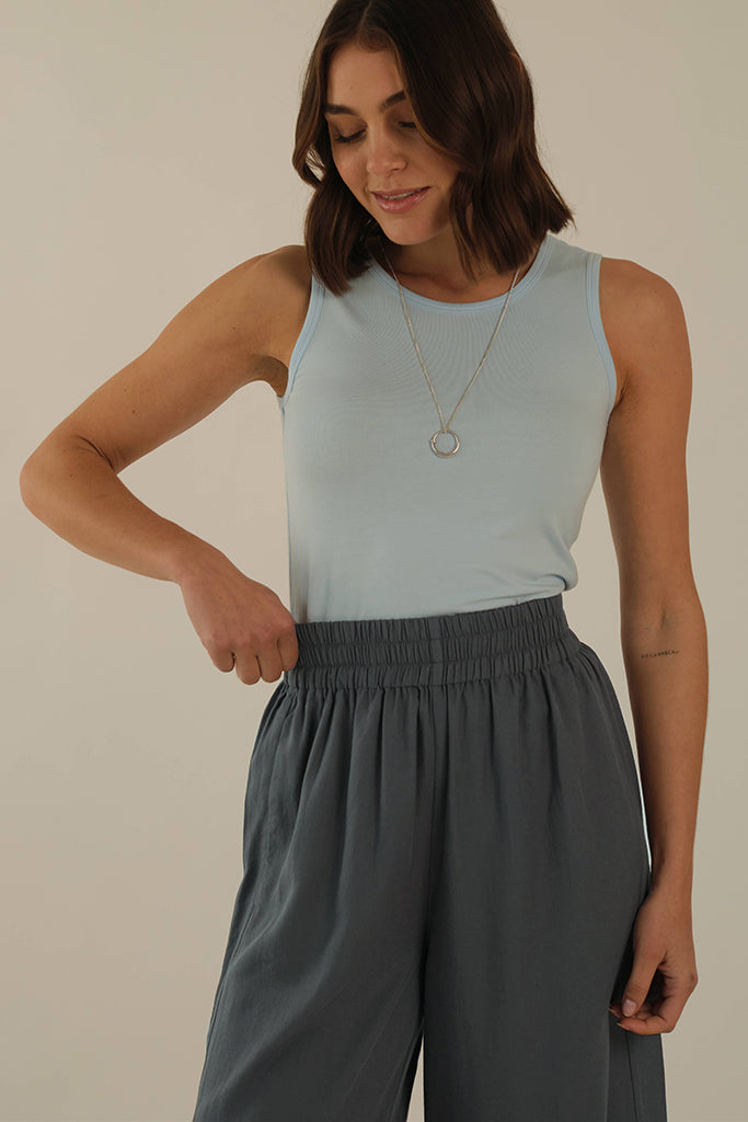 Model is wearing fitted light blue singlet with a long necklace and grey pants