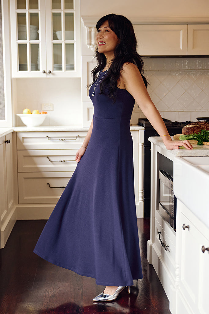 Side view of a woman in a sleeveless indigo-blue maxi dress with wide straps, a fitted bodice, and a long flowing skirt.