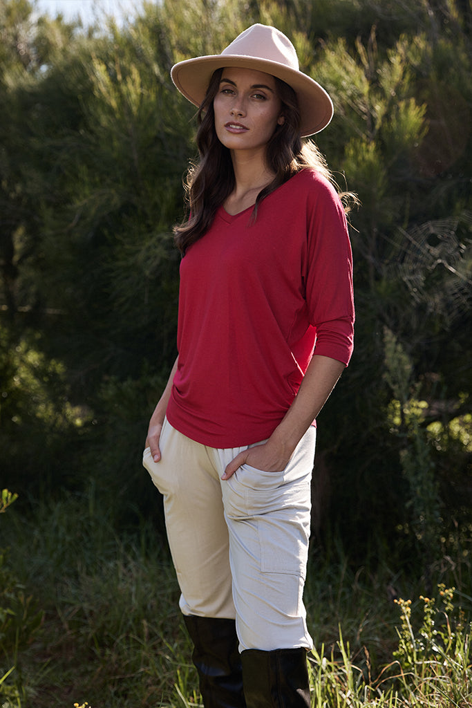 Model standing in a garden wearing a red v neck tee with 3/4 sleeves and beige pants tucked into knee length boots.