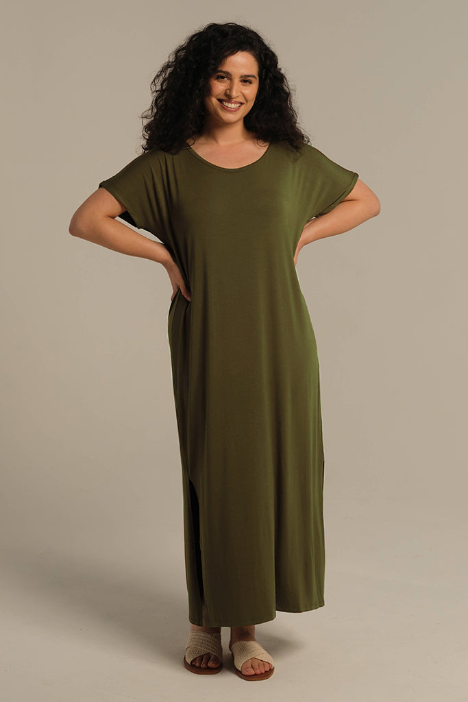 A woman standing in a dark olive maxi dress with a scoop neckline, short sleeves, and side splits to the knee. She is wearing open-toe sandals.