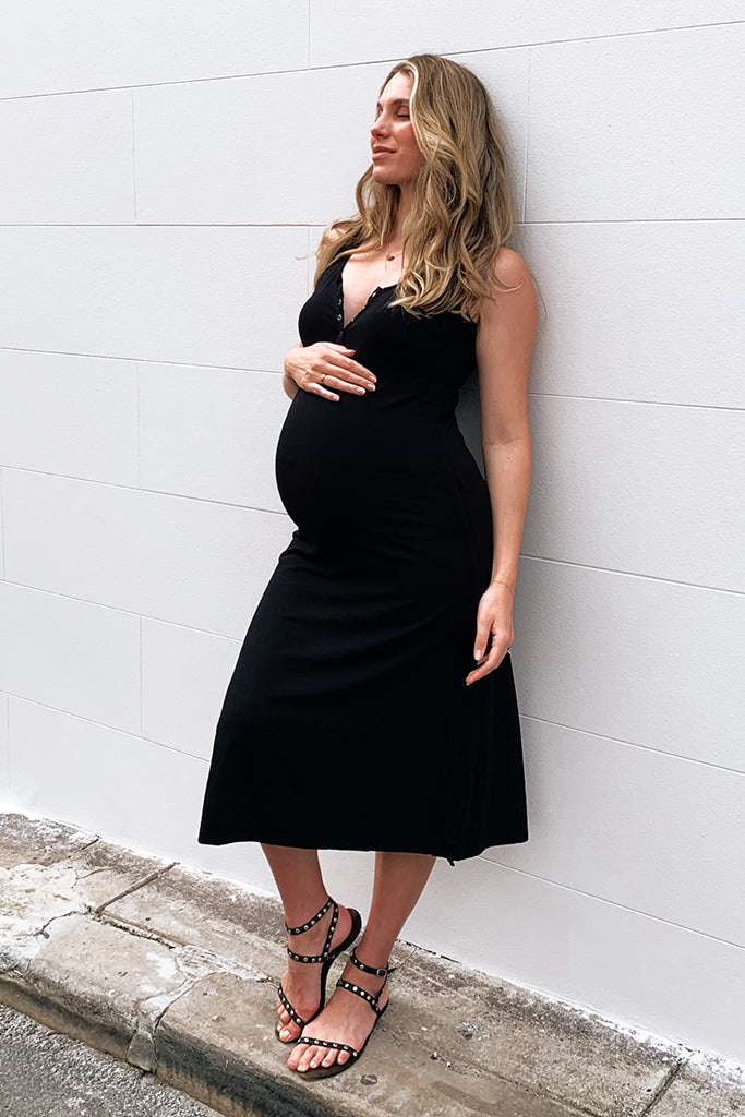 A pregnant woman wearing a black Henley tank maternity dress with small buttons, standing against a white wall.