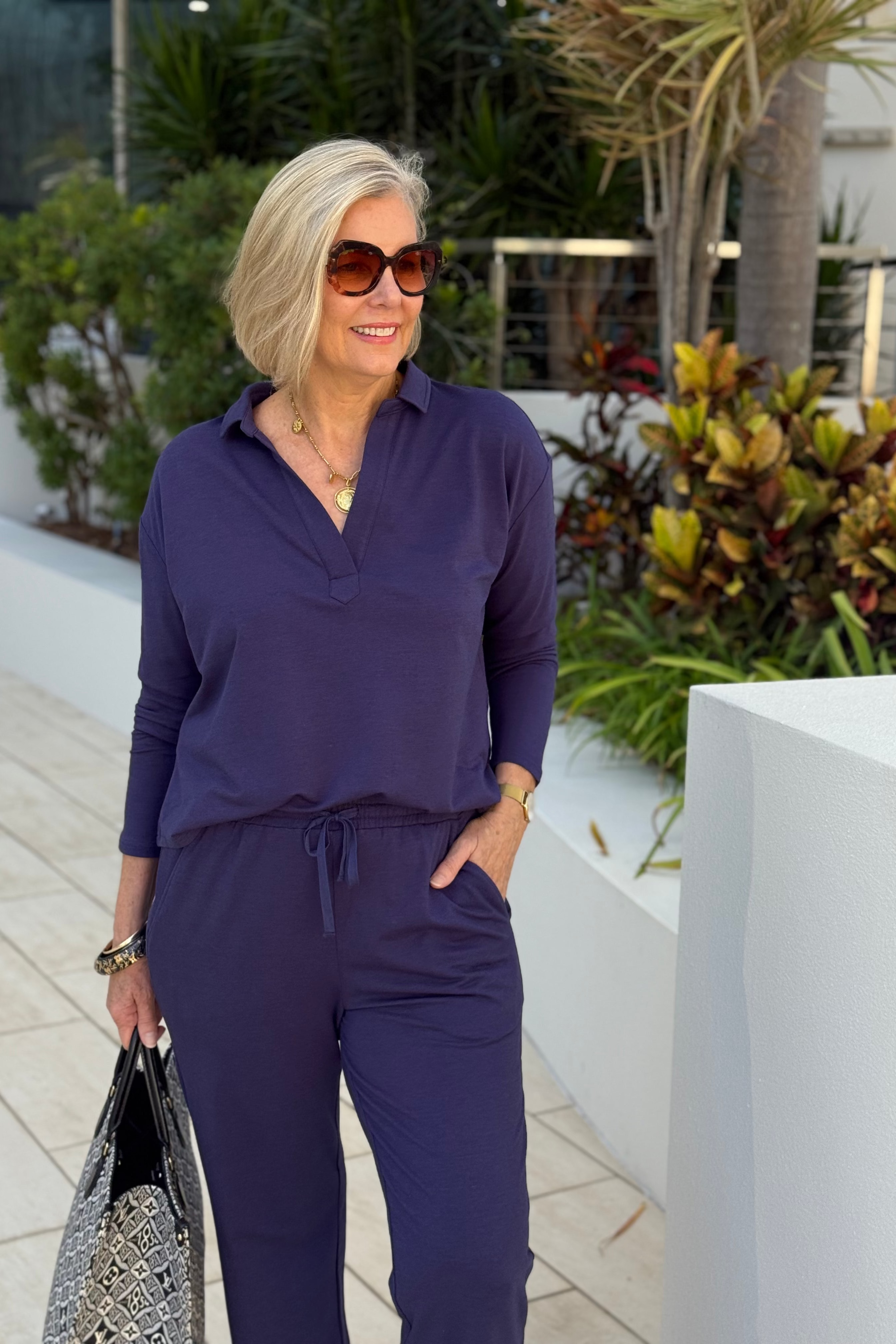 Woman in a navy blue outfit standing outdoors with plants and a building in the background