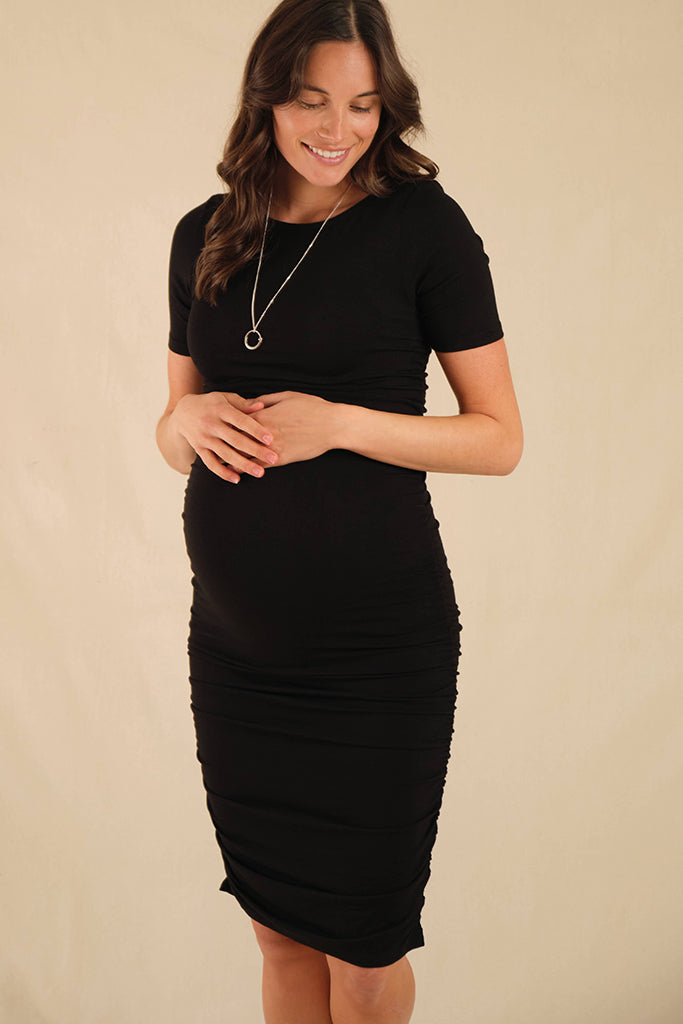 A pregnant woman wearing a short-sleeved, high-necked, black maternity dress with side ruching, standing against a beige background.