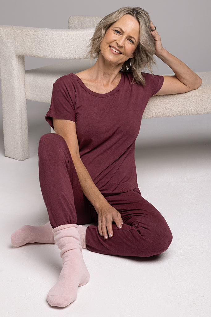 A woman sitting on the floor, wearing a burgundy lounge tee and matching pyjama pants in a relaxed, oversized fit.