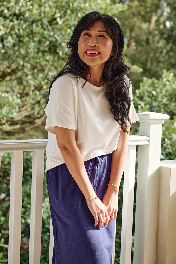Model wearing a white bamboo tee and purple skirt standing on a balcony with greenery in the background