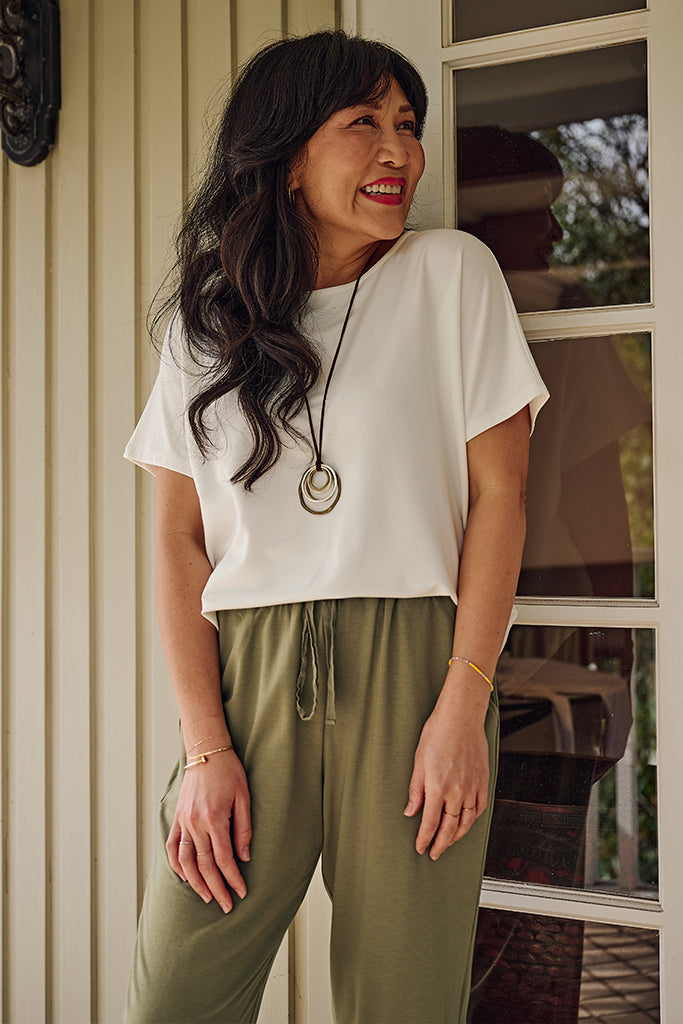Woman wearing a white summer tee and green pants standing in front of a window.