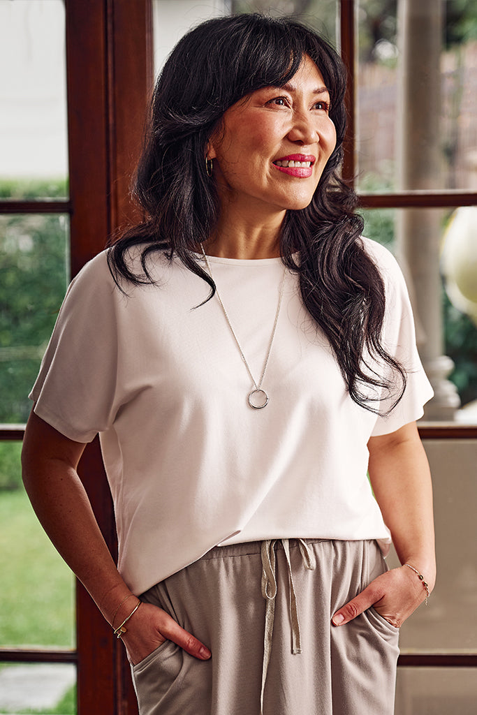 Woman wearing a white short sleeved top and a taupe bamboo skirt standing indoors with a window in the background