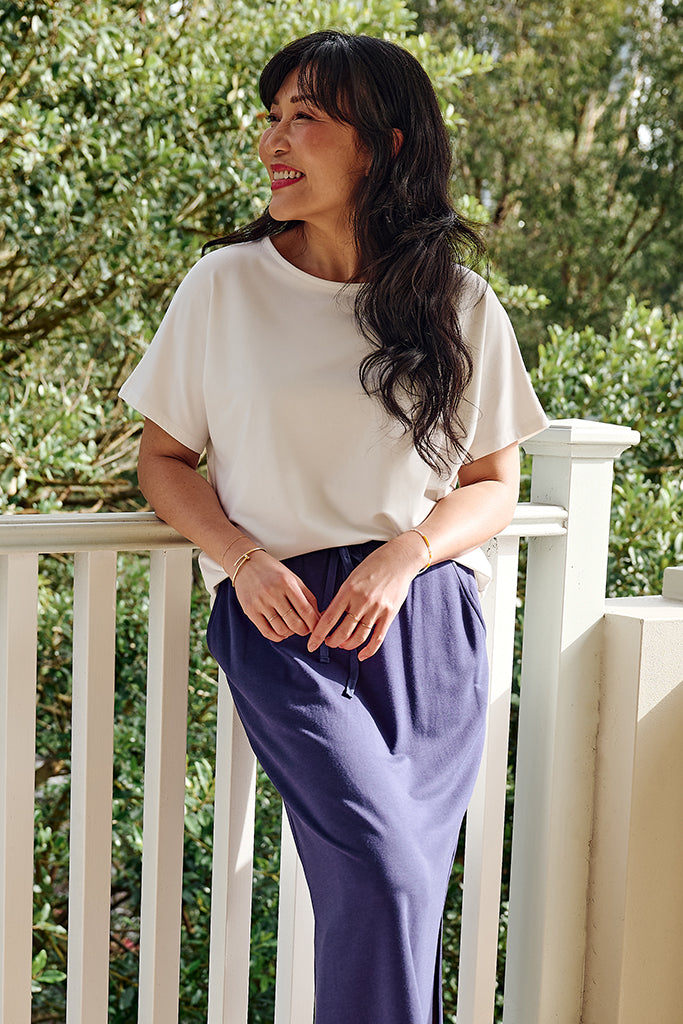 Woman wearing a white short sleeved top and an indigo skirt standing on a balcony with greenery in the background