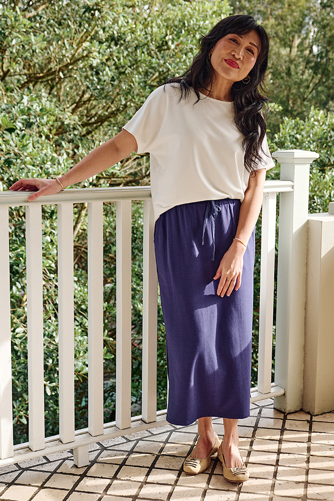 Woman wearing a white tee featuring a high neckline and short sleeves, and a purple skirt on a balcony with greenery in the background