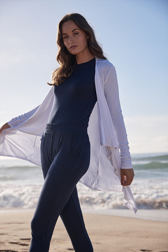 a woman wearing the peggy pants in dark navy with a soft elastic band, side pockets and a tapered leg paired with the same colour layering top and a white waterfall cardigan
