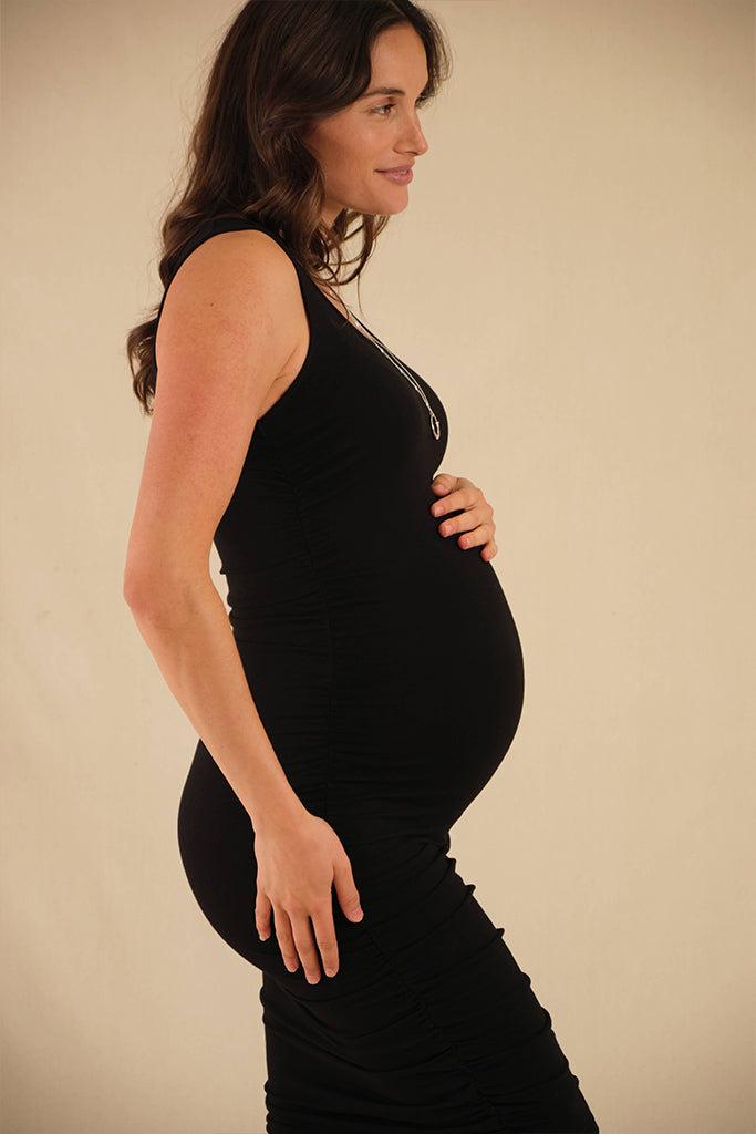 A pregnant woman wearing a black maternity dress with a scoop neckline and ruching down the sides, in a studio setting.
