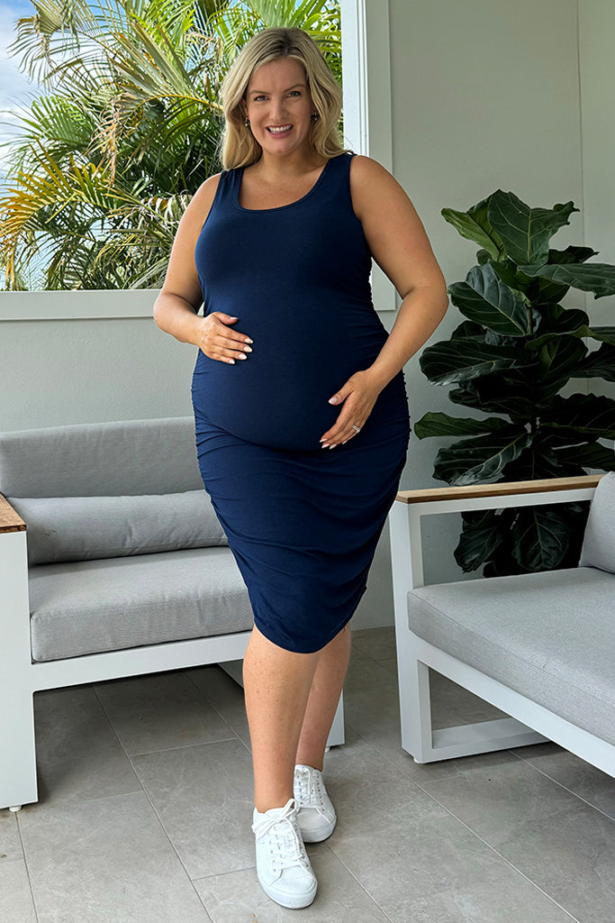 A pregnant woman wearing a navy maternity dress with a scoop neckline and ruching down the sides, paired with white sneakers.