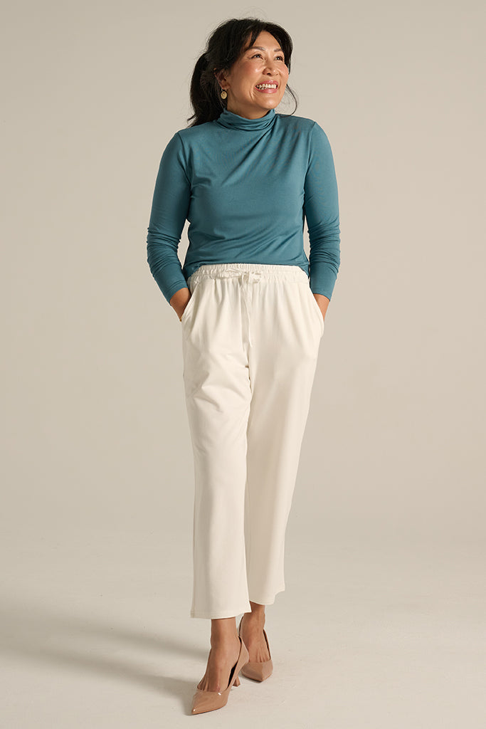 woman wearing ankle length white pants with side pockets paired with the teal turtleneck top