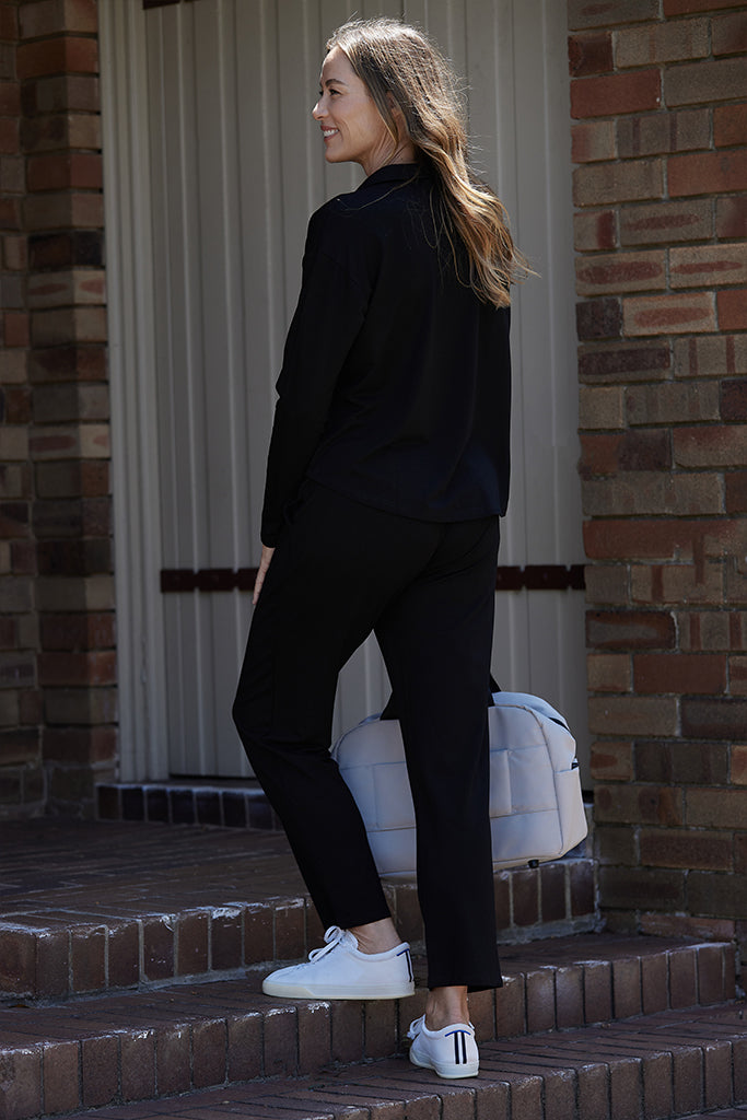 A woman standing on a step wearing black Voyager Pants with an elasticated waistband, side pockets, and a tapered leg design. She is also wearing a black top
