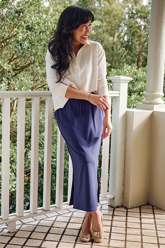 Woman standing on a balcony wearing a white long sleeved tee and purple bamboo skirt.