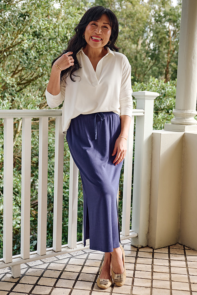 Woman standing on a balcony wearing a white long sleeved top and purple midi skirt.