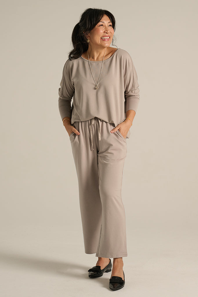 A woman standing wearing taupe-colored straight-leg trousers with an elasticated waistband and side pockets, paired with a matching top.