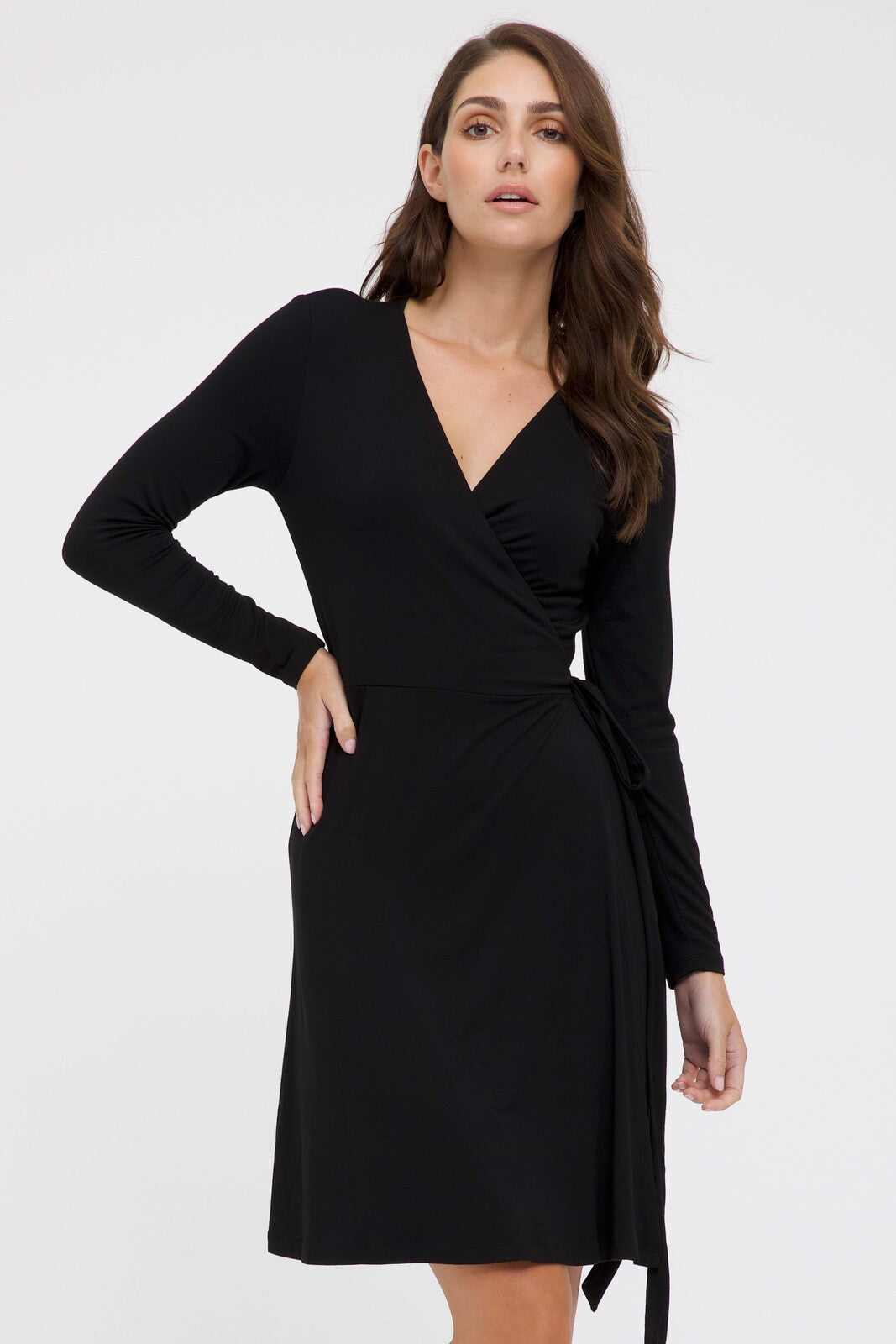 A woman wearing a long sleeve wrap dress in black with a V-neckline and a tie at the waist, standing against a plain background.