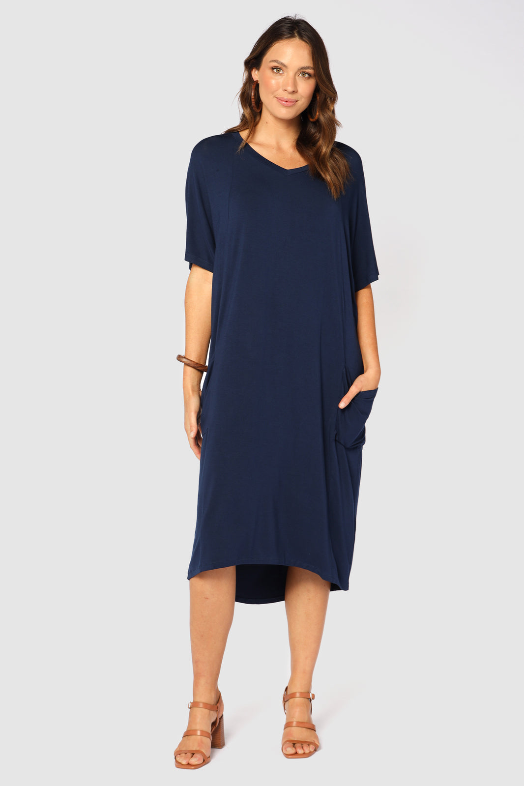 A navy-colored midi length dress with a V neckline, oversized fit, bat-wing sleeves, side pockets, and a hi-lo hem. Model is wearing the dress with a pair of sandals.