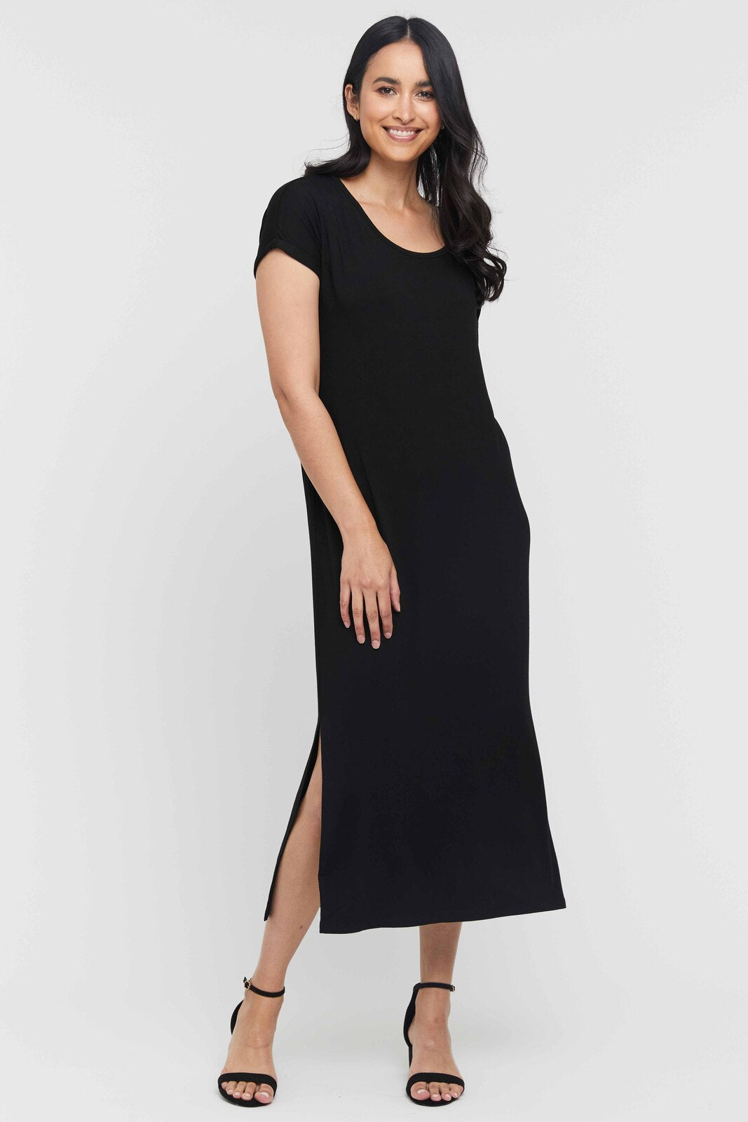 A woman standing in a pose that showcases a black maxi dress with short sleeves, a scoop neckline, and side splits starting at the knee. She is wearing open-toe heels.