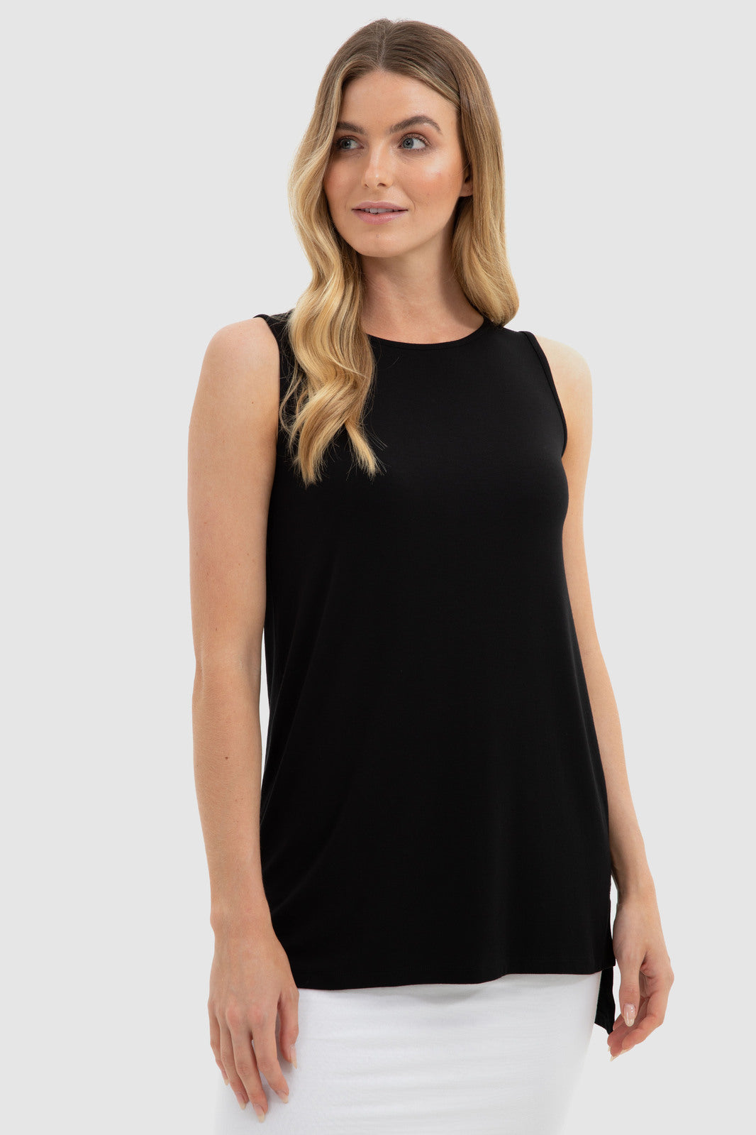 A woman wearing a black sleeveless top with a round neckline, longer back, and side slits. She is paired with white pants.