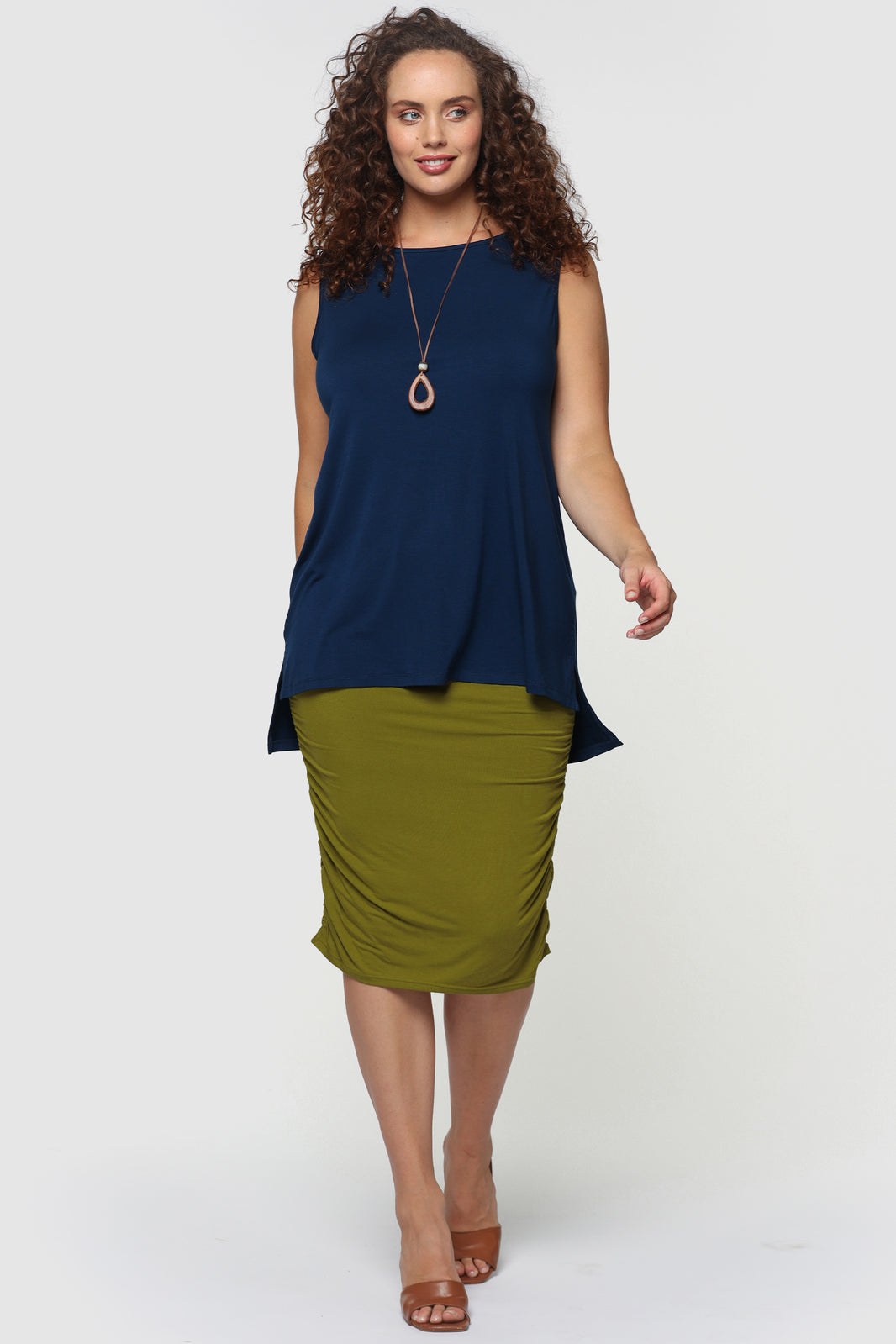 Model is wearing navy blue sleeveless top with high shell neck line pairing it with green skirt and tan sandals.