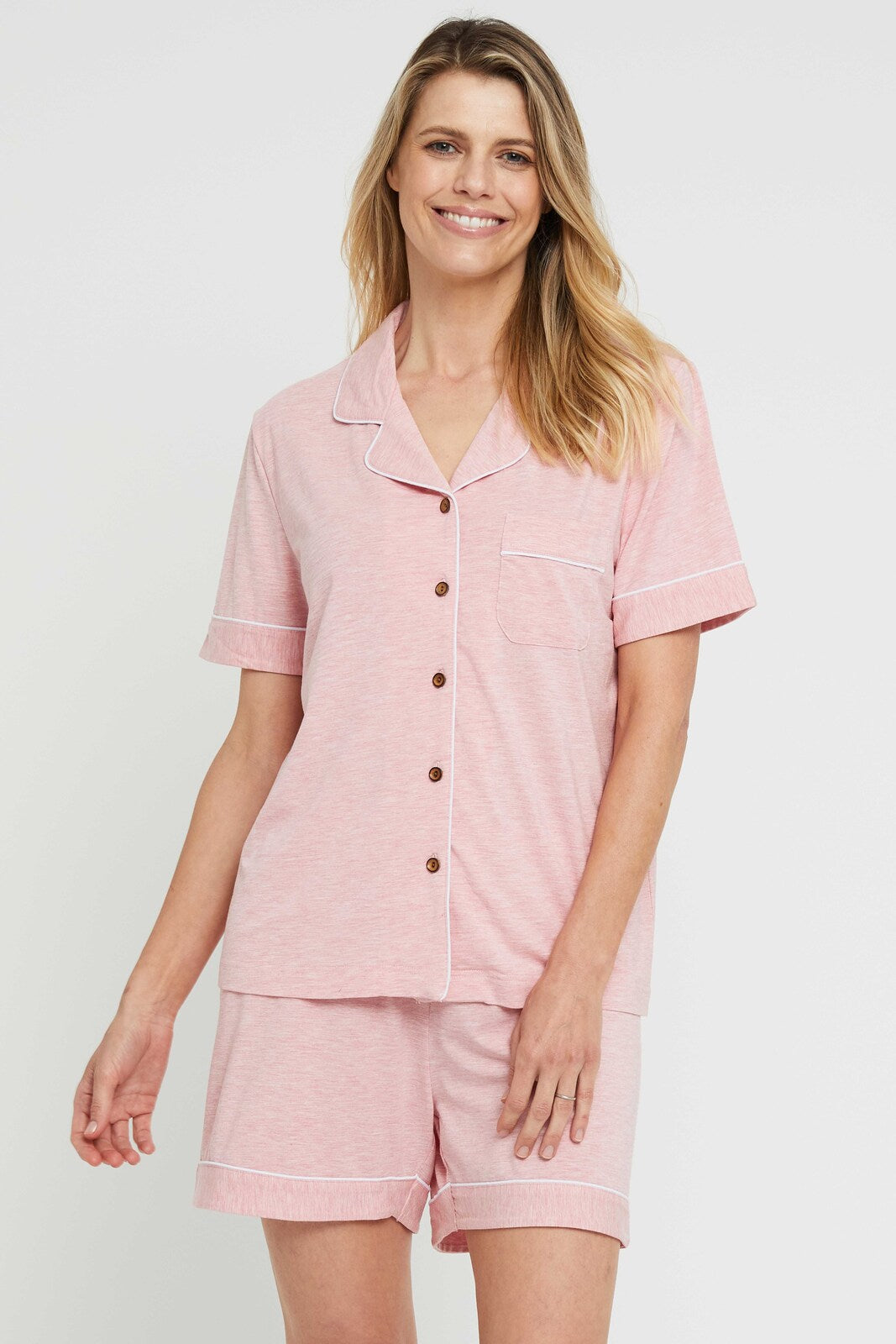 A woman wearing a pink short-sleeved pyjama set with a collar, featuring bamboo buttons and piping details.
