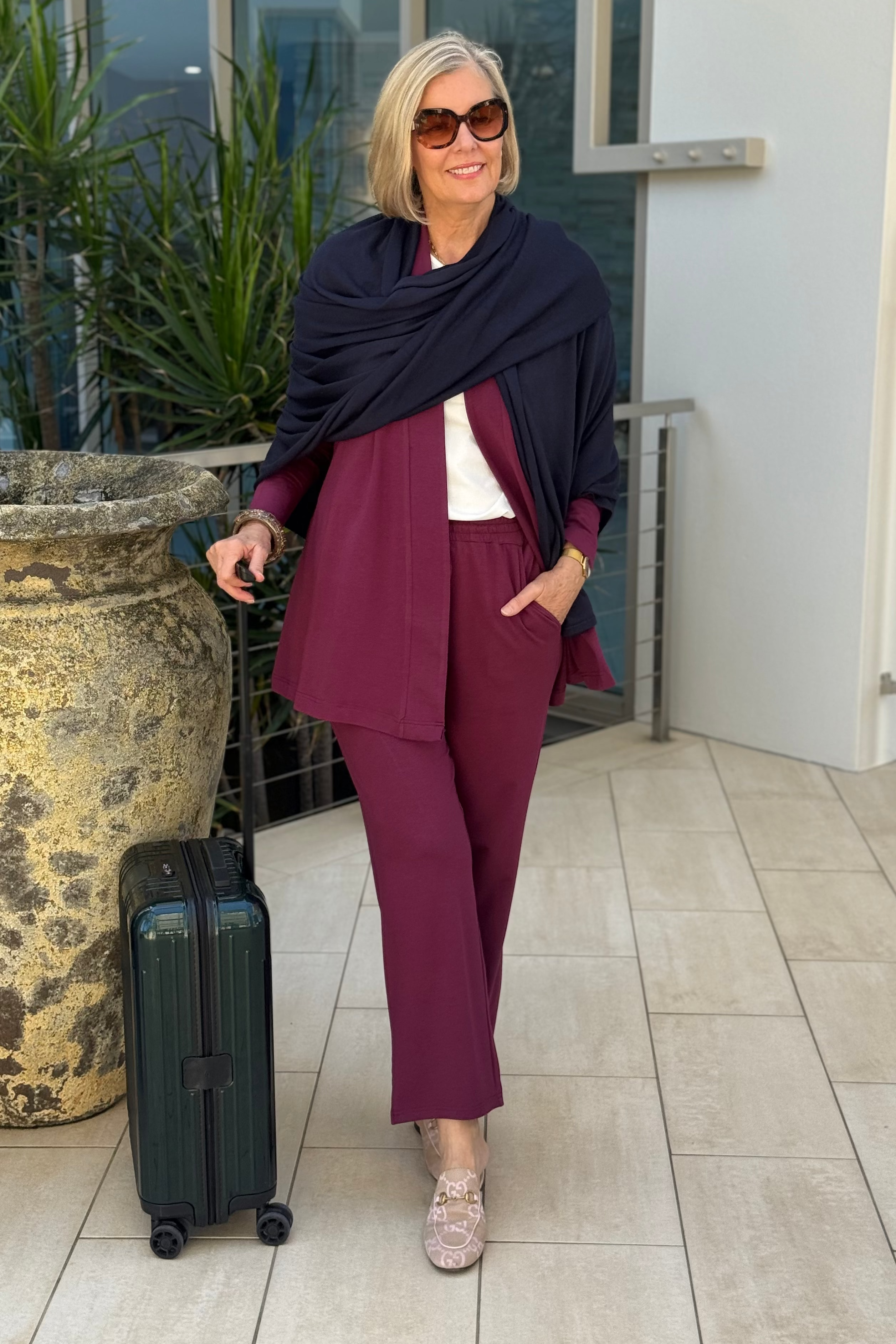 Woman in a burgundy outfit with a navy shawl standing next to a suitcase on a tiled floor.