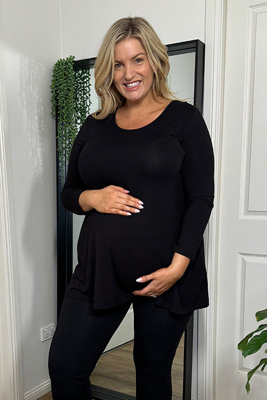 A pregnant woman wearing a black maternity tunic top with long sleeves and a scoop neckline, paired with black leggings and white sneakers.