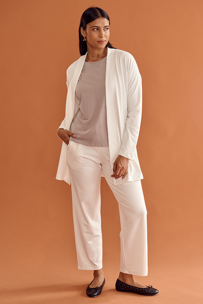 Model wearing a white relaxed cardigan, taupe top, and white pants against an orange background