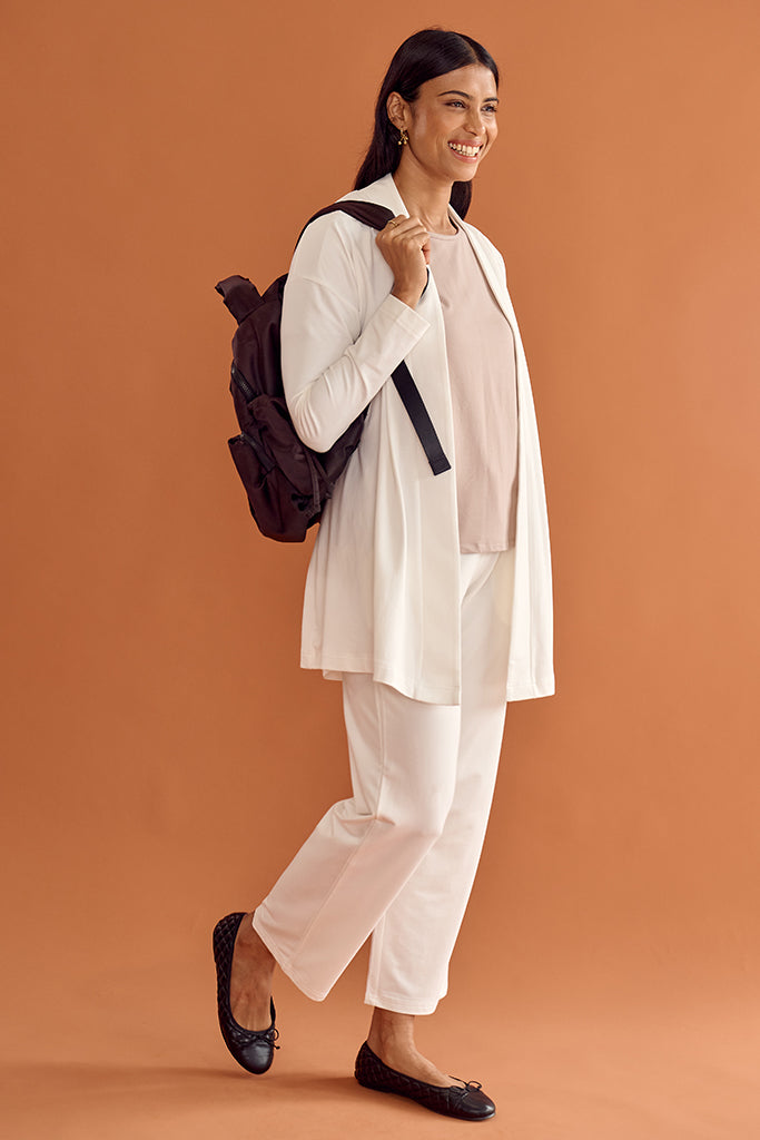 Woman in a white bamboo cardigan and cropped pants holding a black backpack against an orange background